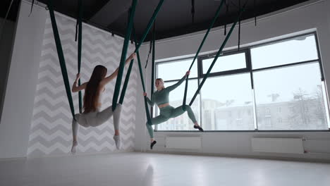 Trainer-and-young-woman-practising-aero-gymnastics-with-suspensions-at-gym.-Group-of-two-young-beautiful-yogi-women-doing-aerial-yoga-practice-in-green-hammocks-in-fitness-club